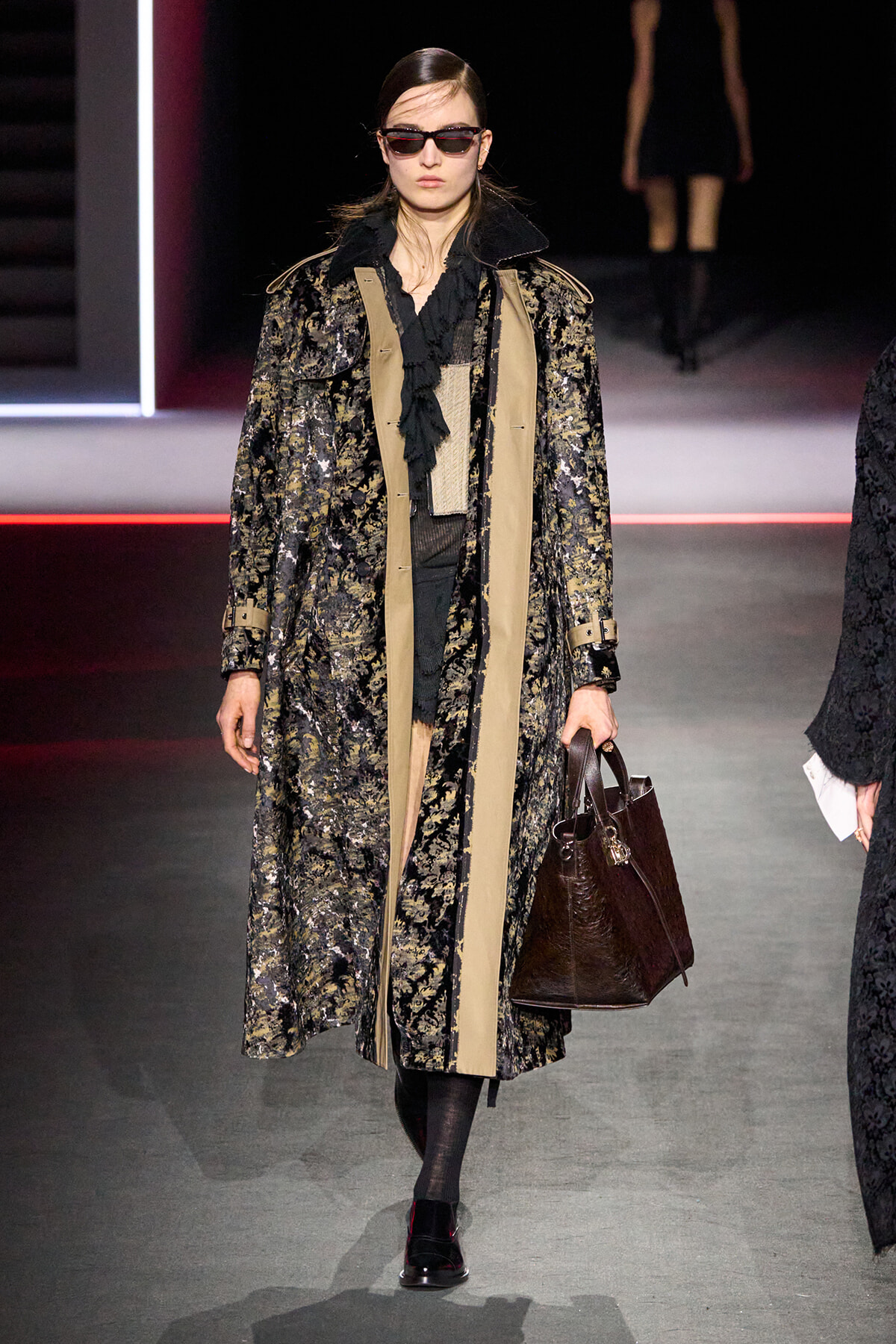 Model walks the runway in a long black-and-gold floral coat with beige panels, black outfit, sunglasses, and a dark brown handbag.