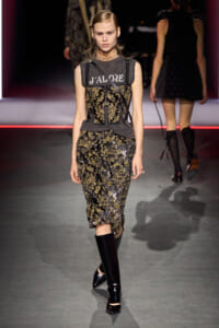 Model walking the runway in a black-and-gold floral lace dress with a gray corset-like overlay and knee-high black boots.