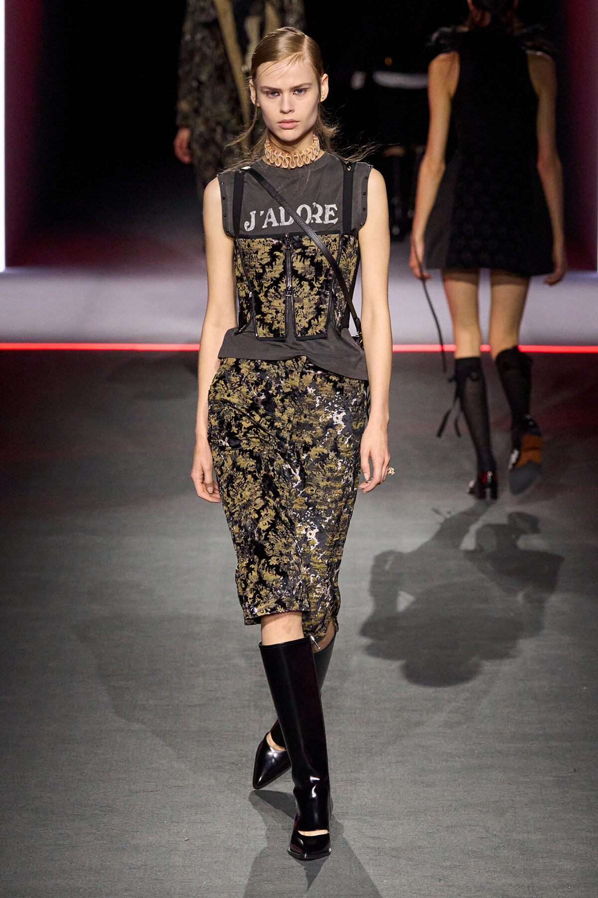 Model walking the runway in a black-and-gold floral lace dress with a gray corset-like overlay and knee-high black boots.