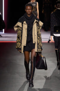 Model on runway wearing a beige and black patterned coat over a black dress, knee-high lace-up boots, and a black handbag.