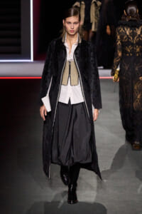 Model walking a fashion show in a long black textured coat over a white shirt, beige corset, and voluminous black trousers with knee-high boots.