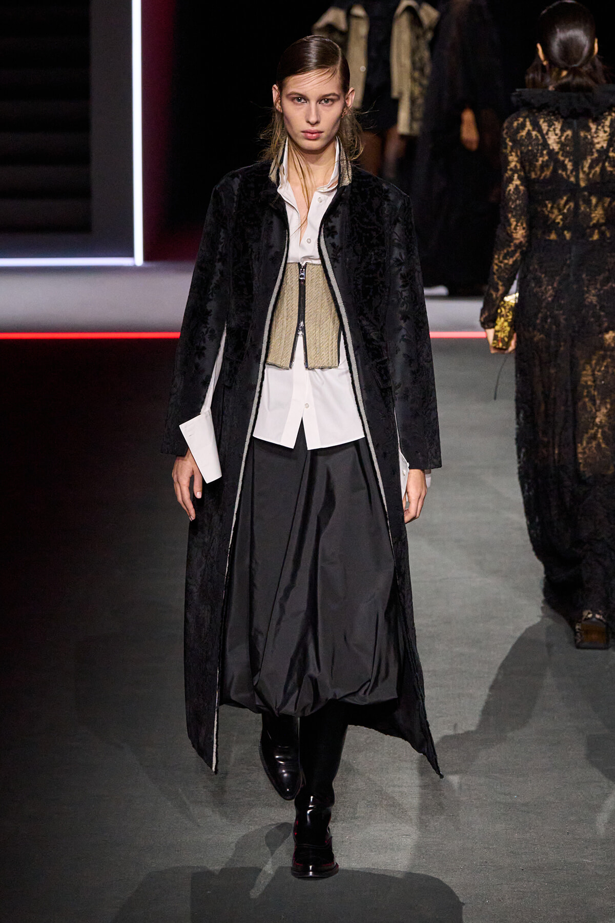 Model walking a fashion show in a long black textured coat over a white shirt, beige corset, and voluminous black trousers with knee-high boots.