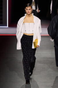 Runway model wearing a white corset top under an open white shirt, black lace pants, a chunky choker, and a gold clutch on a fashion show runway.