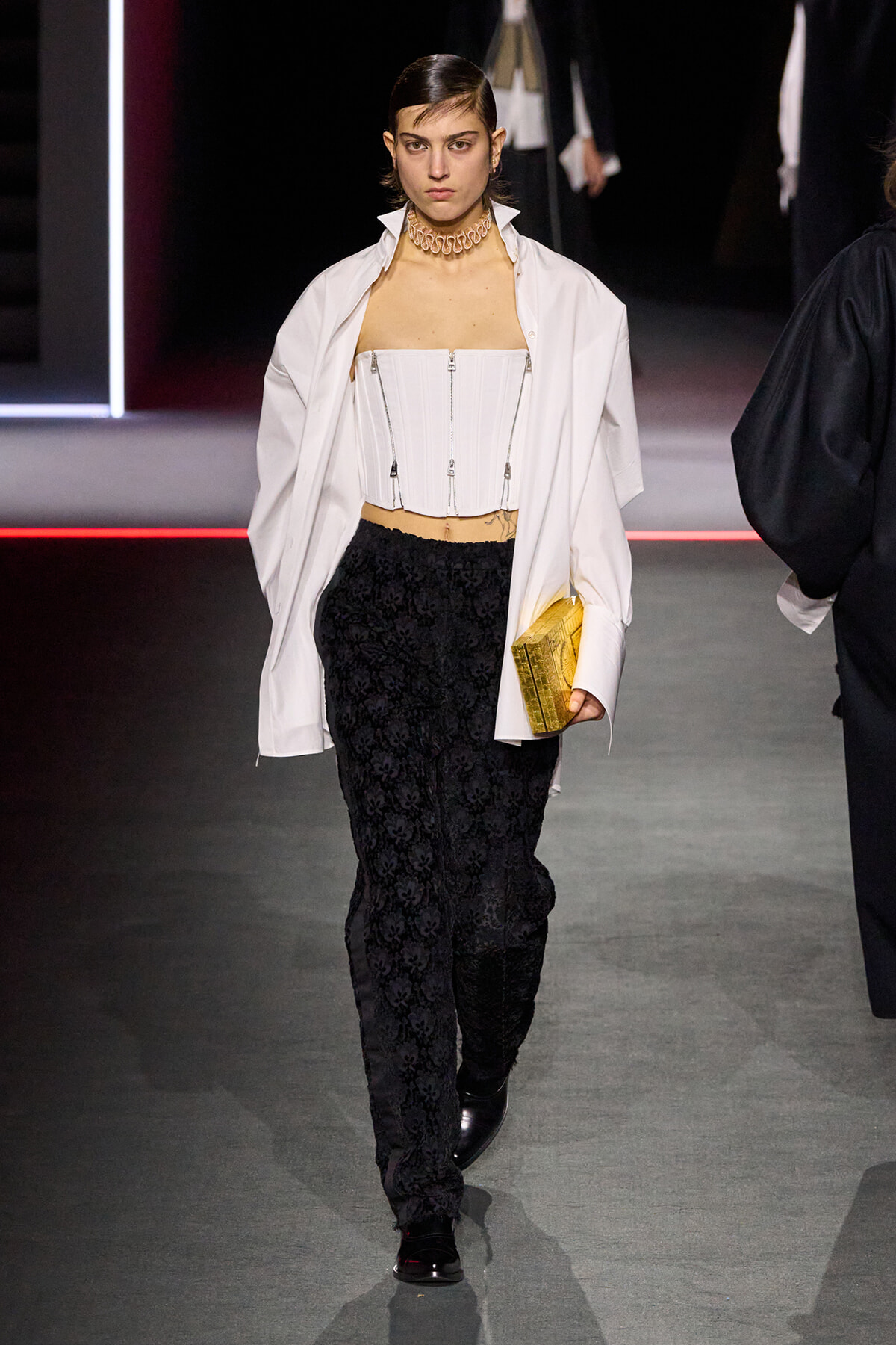 Runway model wearing a white corset top under an open white shirt, black lace pants, a chunky choker, and a gold clutch on a fashion show runway.