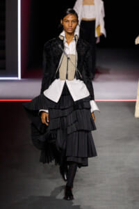 Female model on a runway wearing a white shirt, beige corset belt, black velvet jacket, and layered black pleated skirt.