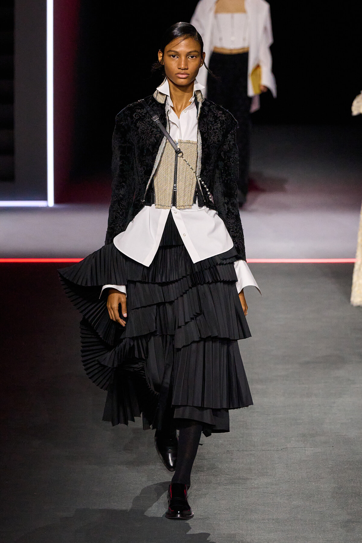 Female model on a runway wearing a white shirt, beige corset belt, black velvet jacket, and layered black pleated skirt.