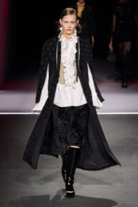 Model on a runway wearing a long black lace coat over a white ruffled blouse, beige corset, and black brocade pants with knee-high boots.