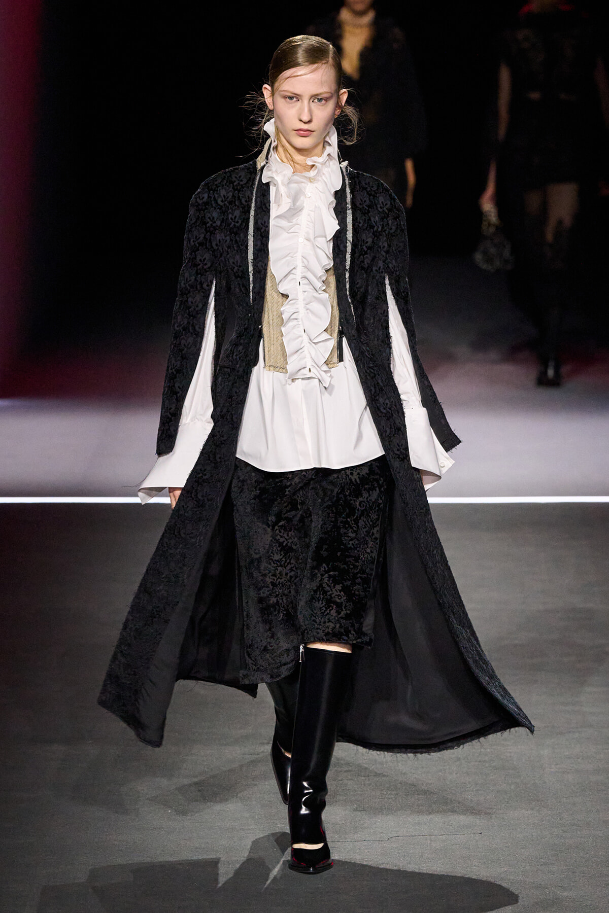 Model on a runway wearing a long black lace coat over a white ruffled blouse, beige corset, and black brocade pants with knee-high boots.