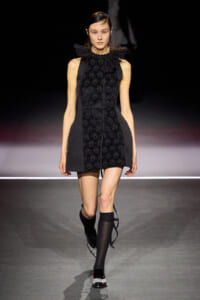 Model walks a runway in a black sleeveless mini dress with a lace front panel, high lace collar, and structured side panels, wearing knee-high socks and embellished shoes.