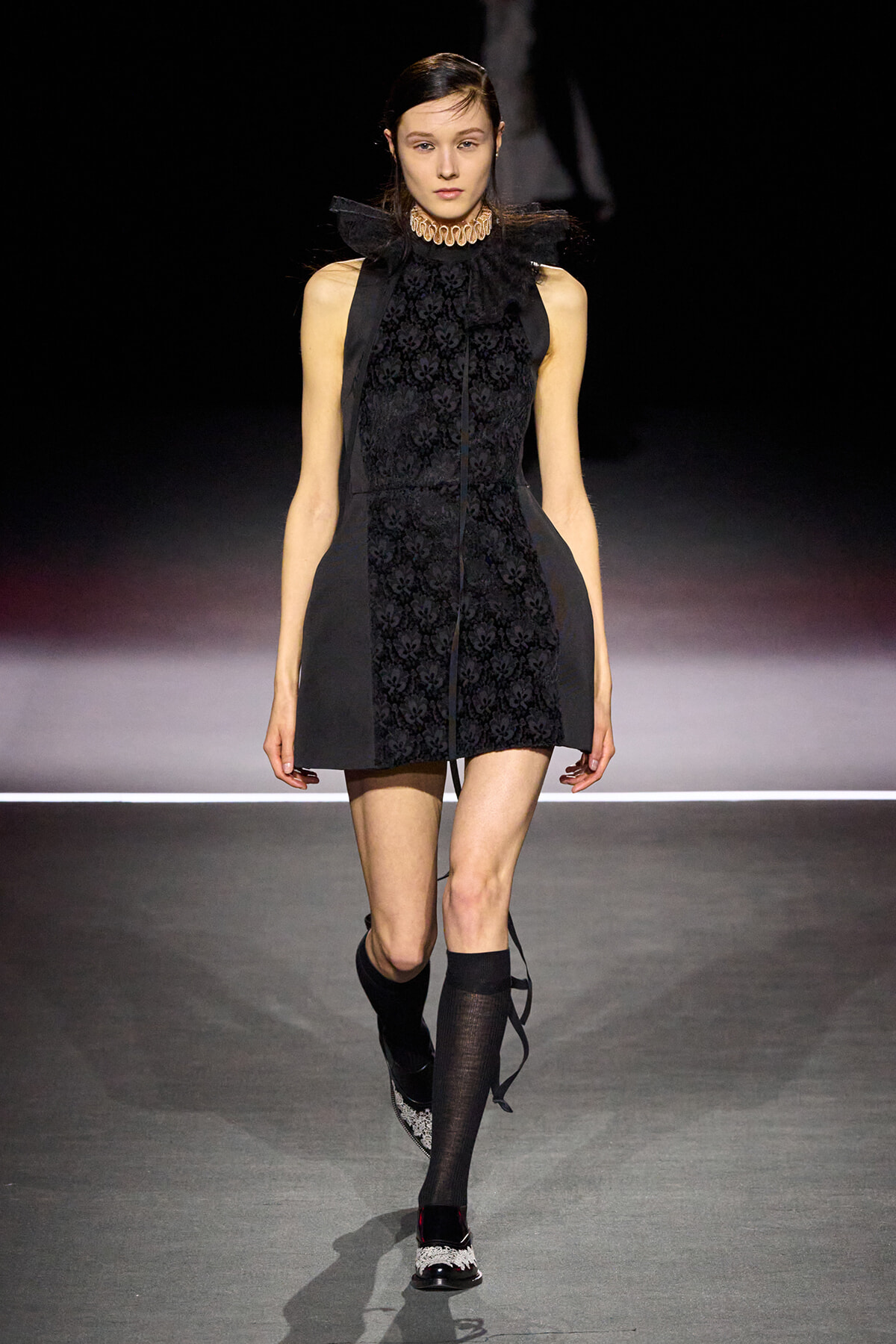 Model walks a runway in a black sleeveless mini dress with a lace front panel, high lace collar, and structured side panels, wearing knee-high socks and embellished shoes.