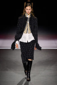 Model walking runway in a black textured suit with a white shirt and beige vest, paired with knee-high black boots and a matching black jacket with ruffle details