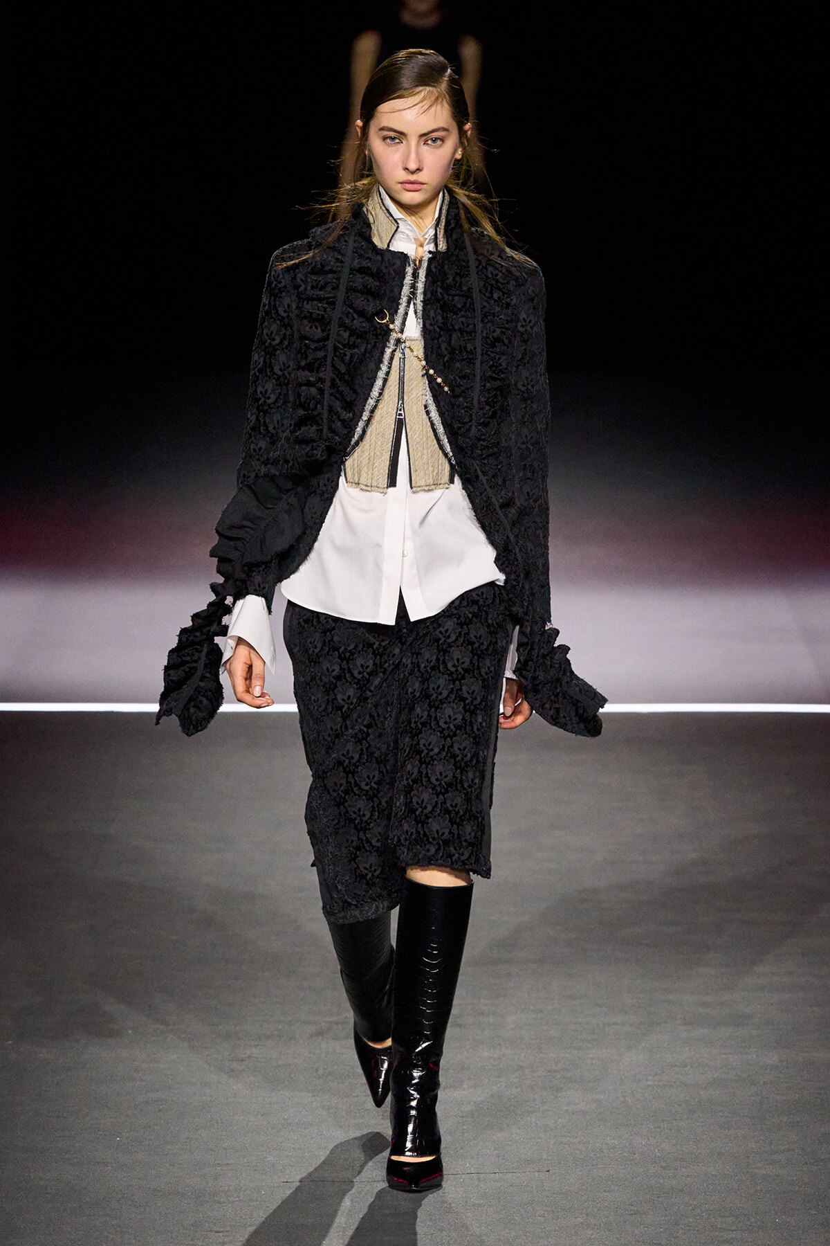 Model walking runway in a black textured suit with a white shirt and beige vest, paired with knee-high black boots and a matching black jacket with ruffle details