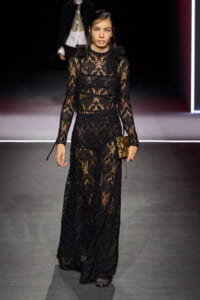 Model walking a runway in a full-length black lace gown with sheer panels and a ruffled high neck, carrying a small patterned clutch.
