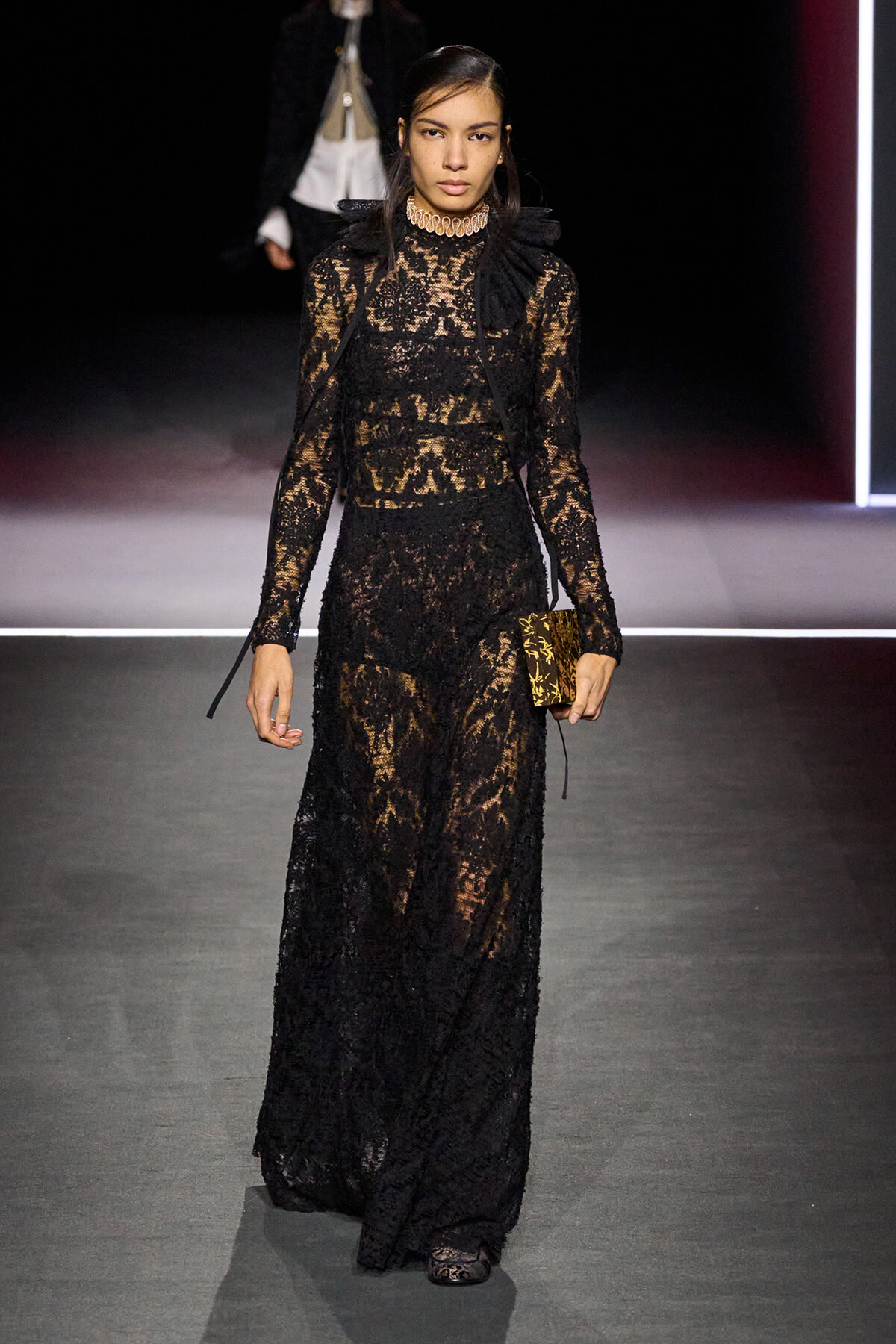 Model walking a runway in a full-length black lace gown with sheer panels and a ruffled high neck, carrying a small patterned clutch.
