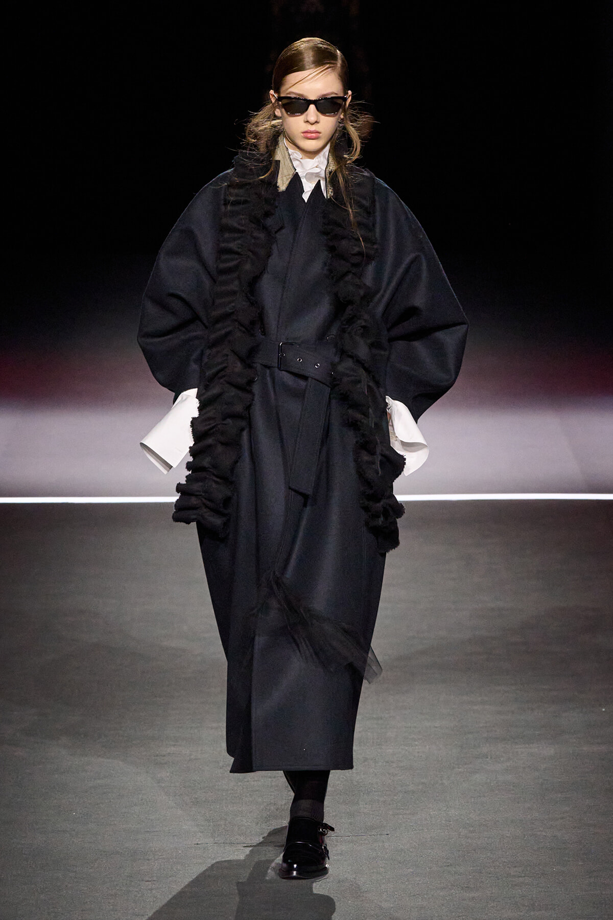 Model walking a runway in an oversized black coat with ruffled trim, white shirt cuffs, and a belt, wearing dark sunglasses.
