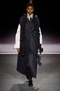 Model walks the runway in a long dark coat with dramatic black ruffles over white oversized sleeves.
