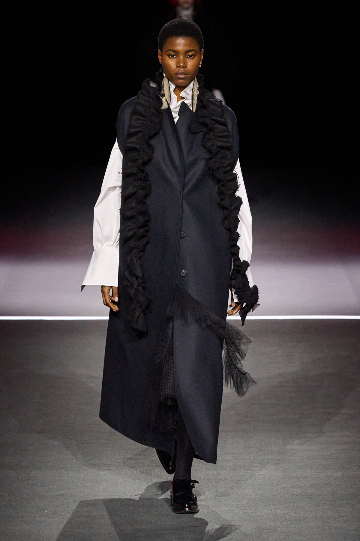 Model walks the runway in a long dark coat with dramatic black ruffles over white oversized sleeves.