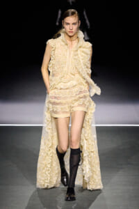 Model on a fashion runway wearing a cream lace outfit with layered ruffles, sheer long cape, and black knee-high socks with loafers.