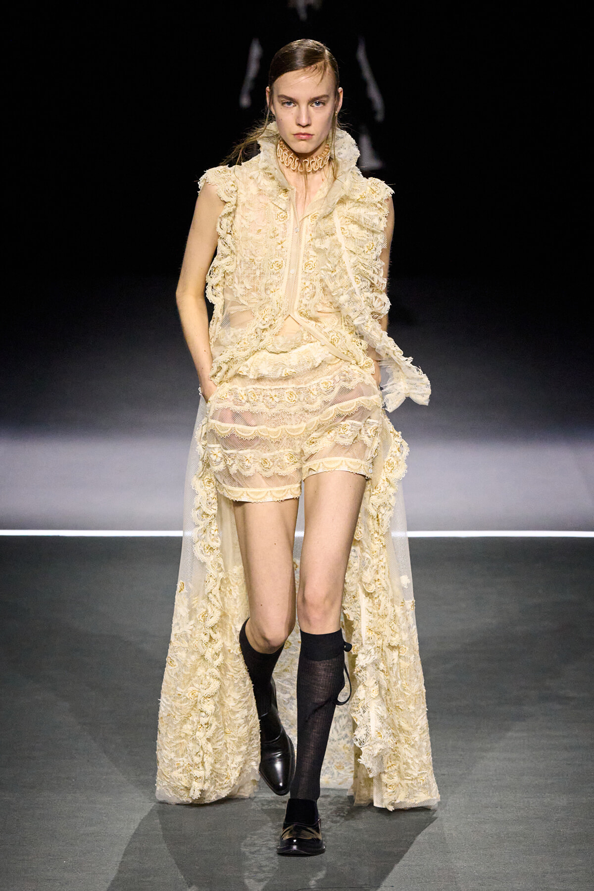 Model on a fashion runway wearing a cream lace outfit with layered ruffles, sheer long cape, and black knee-high socks with loafers.
