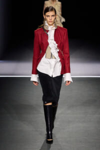 Model walks a runway in a burgundy red blazer over a white ruffled shirt, beige corset, black pants, and tall glossy black boots.