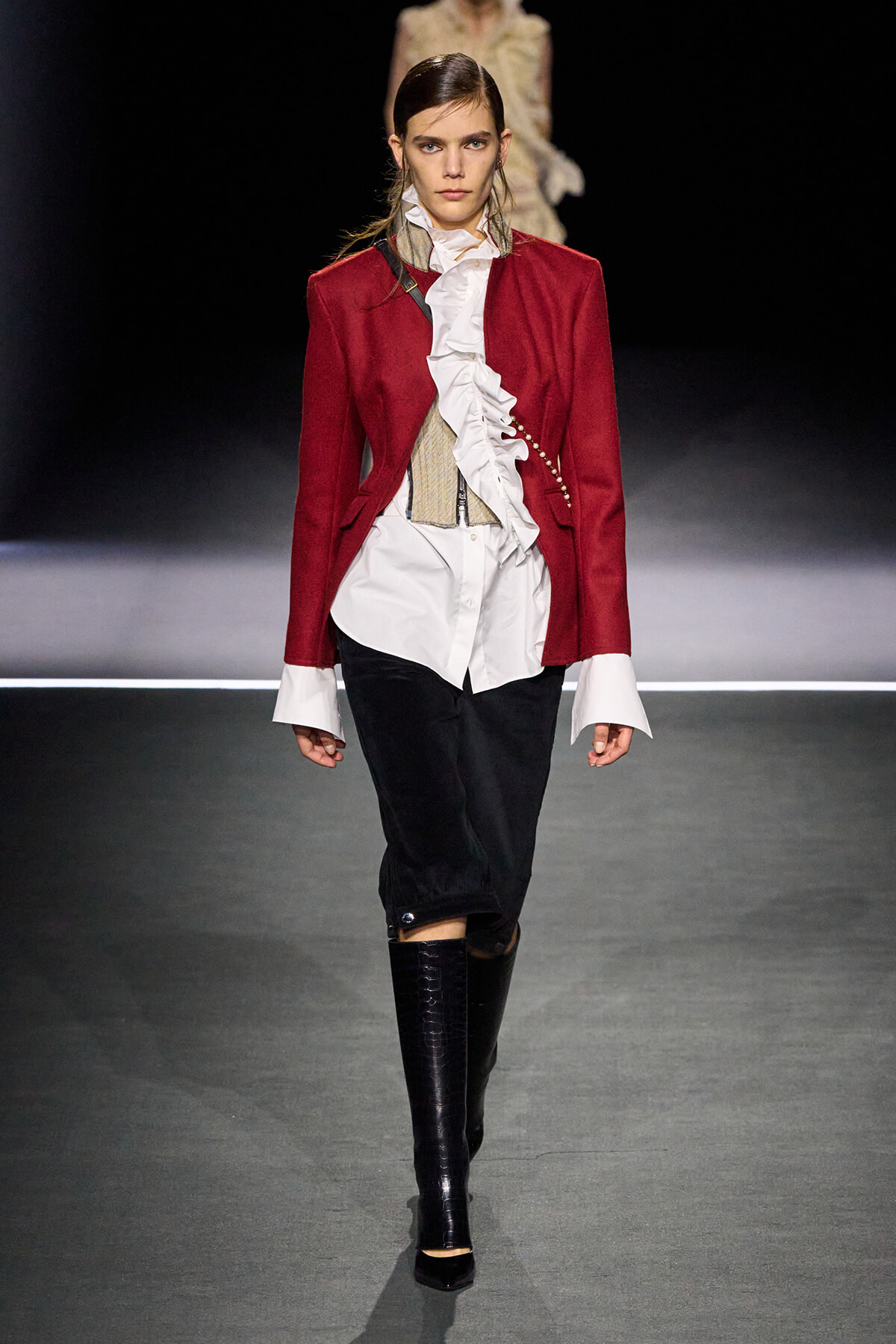 Model walks a runway in a burgundy red blazer over a white ruffled shirt, beige corset, black pants, and tall glossy black boots.