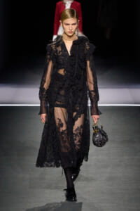 Model walks the runway in a sheer black lace dress with ruffles and long sleeves, carrying a small black handbag with a chain handle.