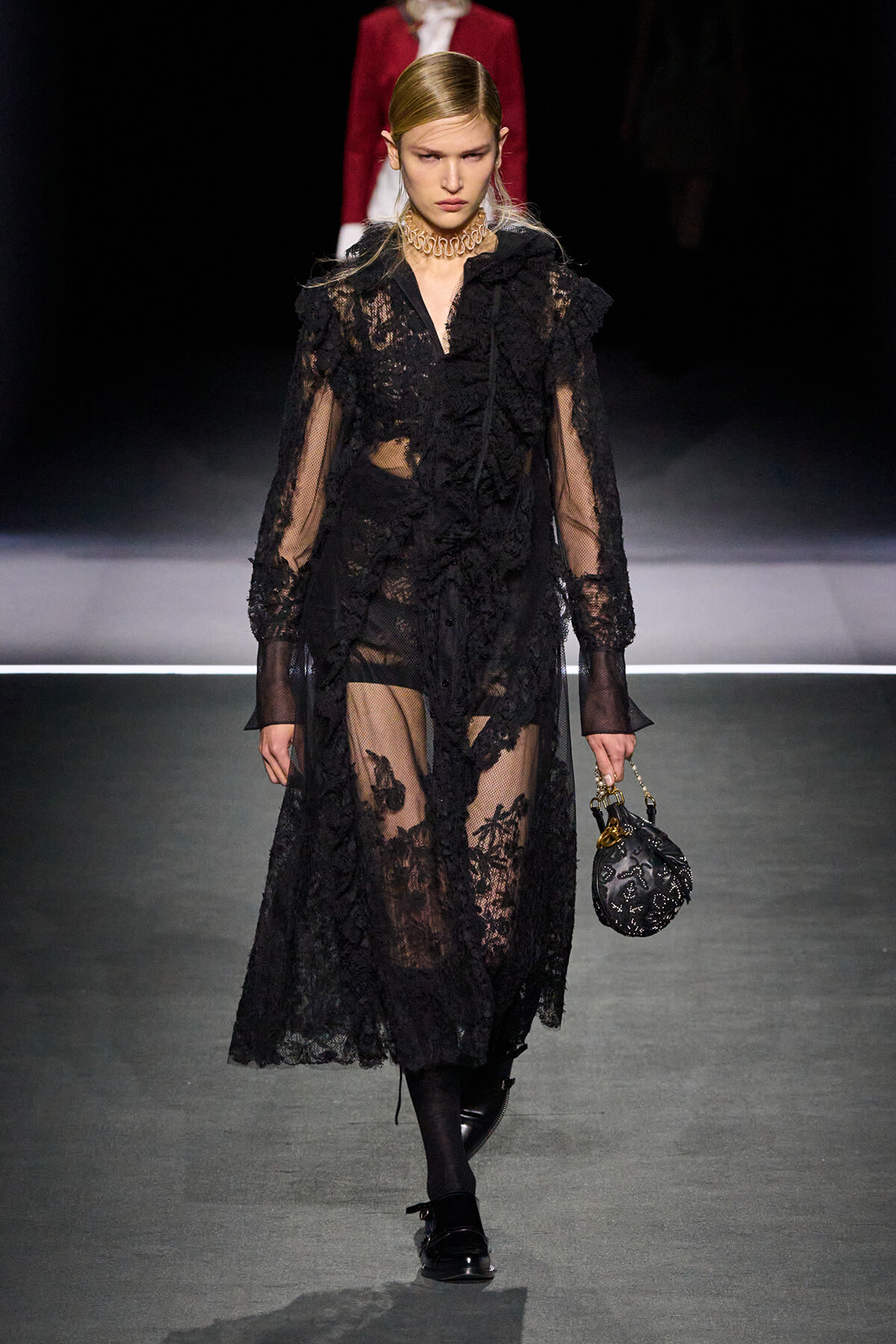 Model walks the runway in a sheer black lace dress with ruffles and long sleeves, carrying a small black handbag with a chain handle.