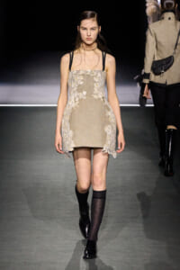 Model walking the runway in a beige lace mini dress with floral appliqués, black straps, and a choker necklace.