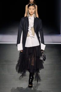 Model walks runway in a dark blazer over a white ruffled blouse, beige corset, and tiered black tulle skirt, holding a small black handbag.