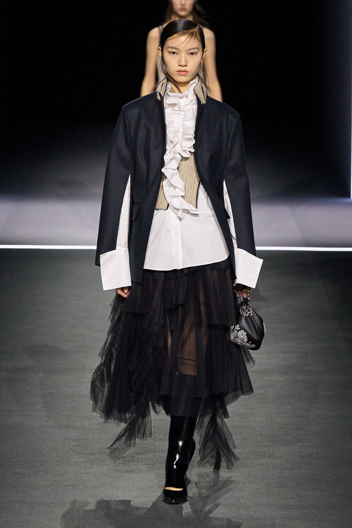 Model walks runway in a dark blazer over a white ruffled blouse, beige corset, and tiered black tulle skirt, holding a small black handbag.