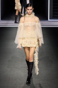 Runway model in a cream lace mini dress with a sheer cape and long lace train, on a fashion show runway.
