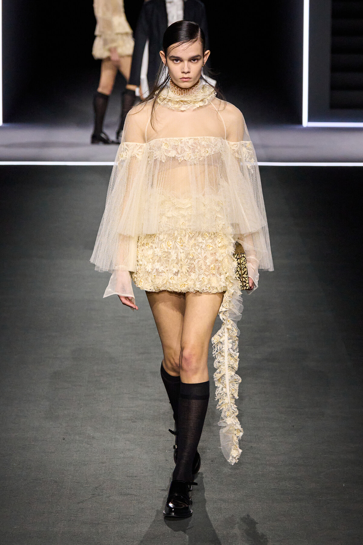 Runway model in a cream lace mini dress with a sheer cape and long lace train, on a fashion show runway.
