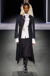 Model walks a fashion runway wearing a black long coat over a white ruffled shirt, paired with a layered black skirt and embellished shoes.