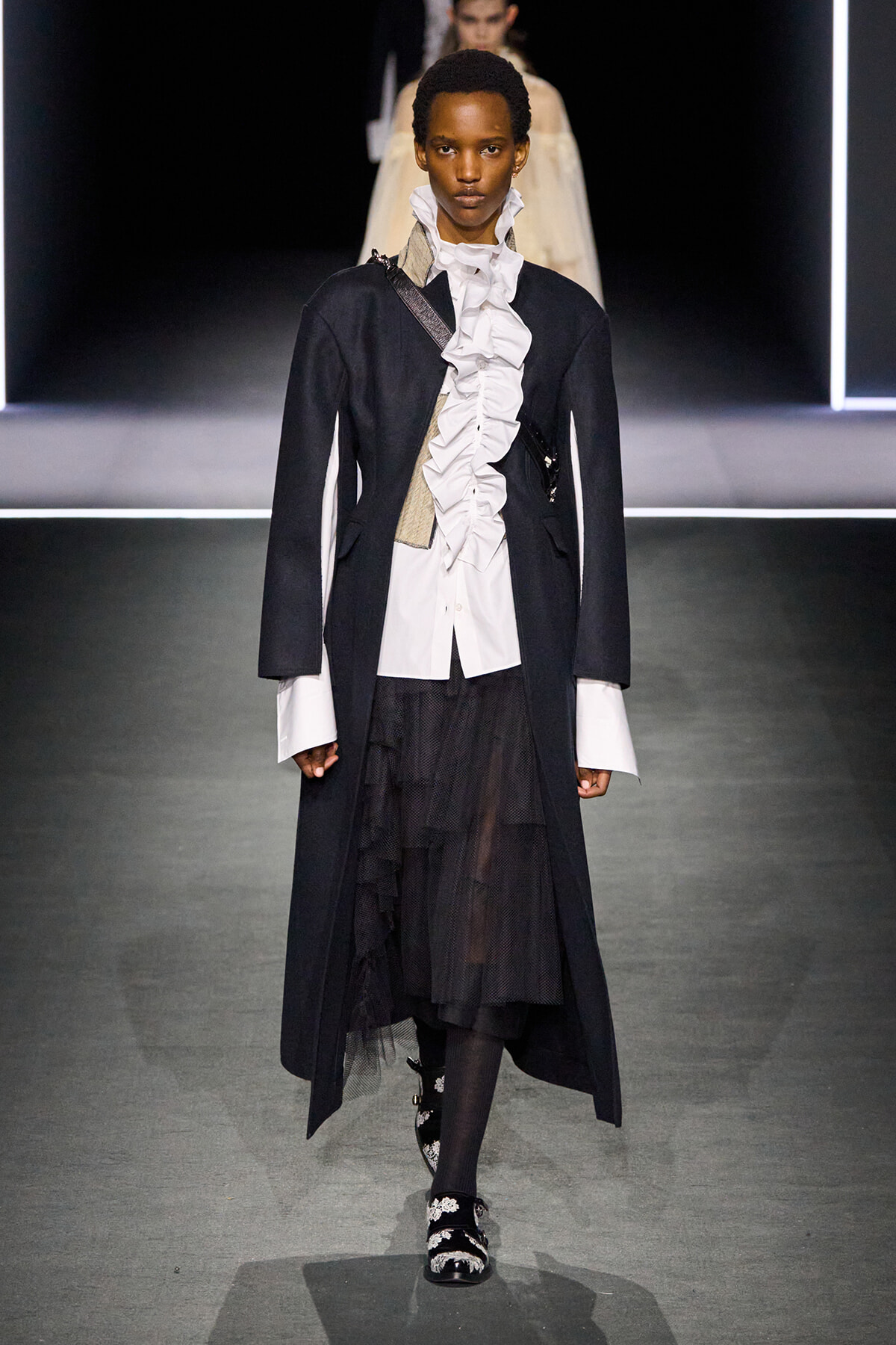 Model walks a fashion runway wearing a black long coat over a white ruffled shirt, paired with a layered black skirt and embellished shoes.