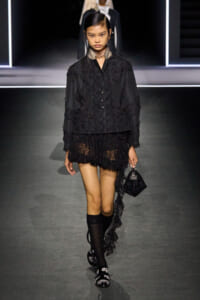 Model on a runway wearing a black lace jacket and tiered mini skirt, carrying a small black handbag; knee-high socks and embellished sandals complete the look.
