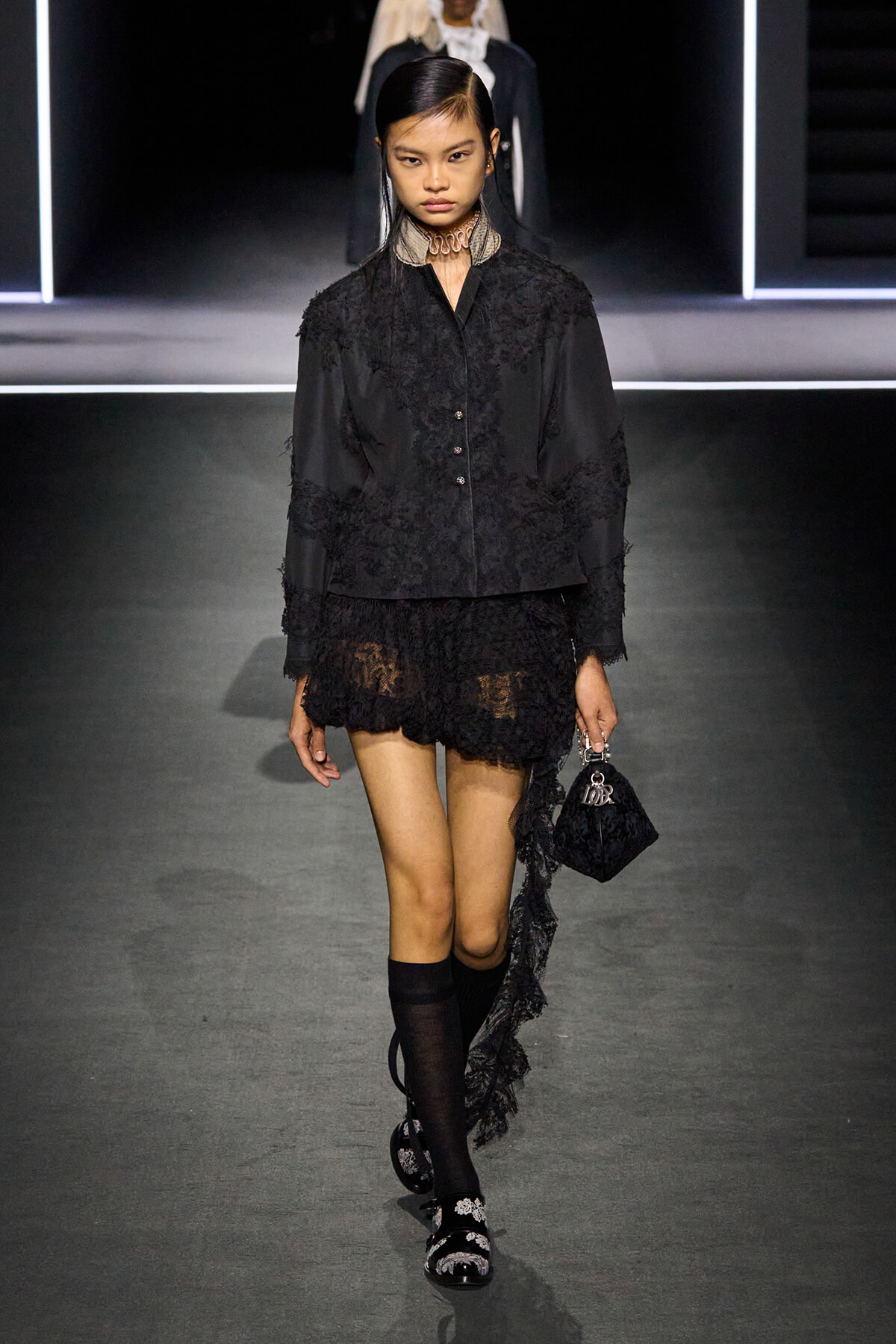 Model on a runway wearing a black lace jacket and tiered mini skirt, carrying a small black handbag; knee-high socks and embellished sandals complete the look.