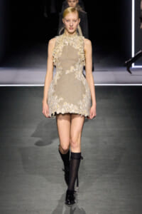 Model walking a fashion runway in a beige lace mini dress with floral appliqués and high neck, black knee-high socks, and black shoes