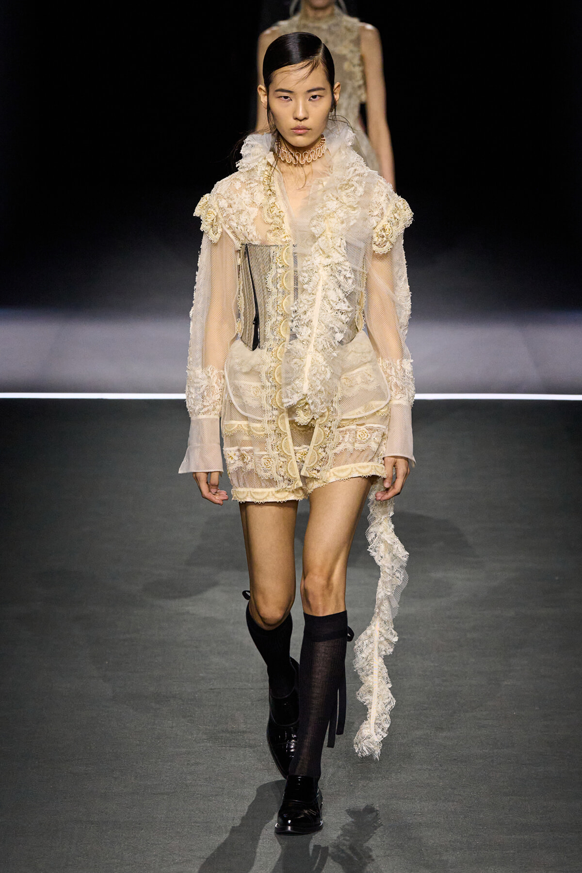 Model walking a runway in a vintage ivory lace ensemble with sheer sleeves and ruffled scarf accessory.