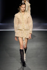 Runway model in ivory lace ensemble: high-neck jacket with floral appliqué and a tiered lace mini skirt, with a long lace train down one leg and black knee-high socks.