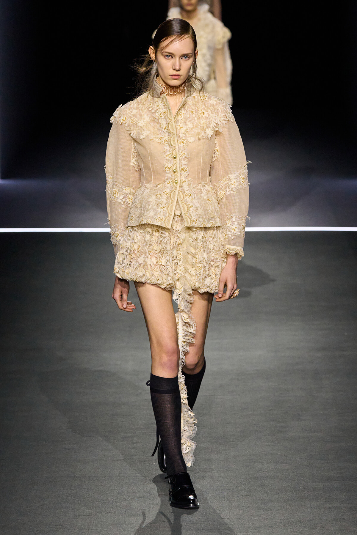 Runway model in ivory lace ensemble: high-neck jacket with floral appliqué and a tiered lace mini skirt, with a long lace train down one leg and black knee-high socks.