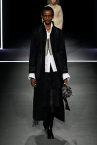 Model on a runway wearing a long black coat over a white shirt, layered black skirt, and a small textured handbag.