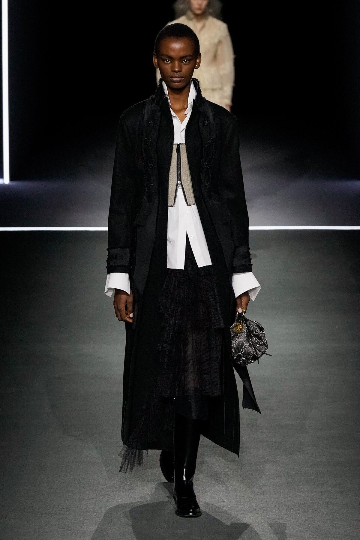 Model on a runway wearing a long black coat over a white shirt, layered black skirt, and a small textured handbag.
