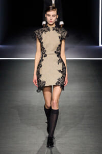 Model walks the runway wearing a beige sheath dress with black floral lace appliqués along the edges and sleeves, creating a contrast look.