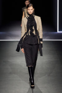 Model walking runway in a beige textured blazer over a black ruffled blouse and black knee-length pants, paired with shiny black knee-high boots.
