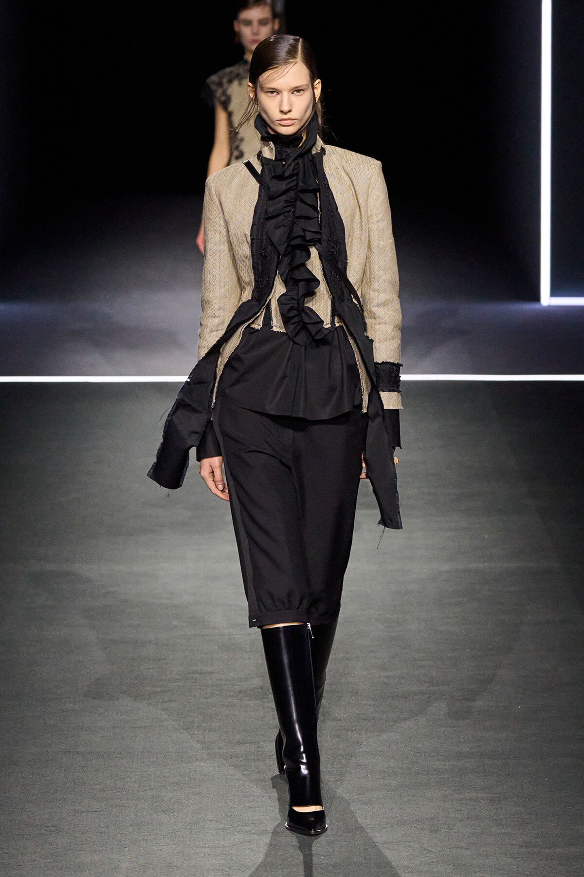Model walking runway in a beige textured blazer over a black ruffled blouse and black knee-length pants, paired with shiny black knee-high boots.