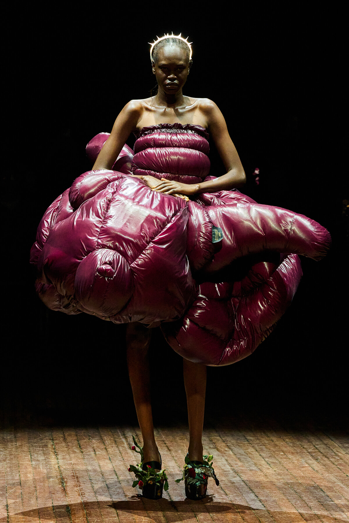 Model on a runway wearing an oversized magenta puffer gown with a halo-like headpiece; dramatic fashion statement on a dark stage.