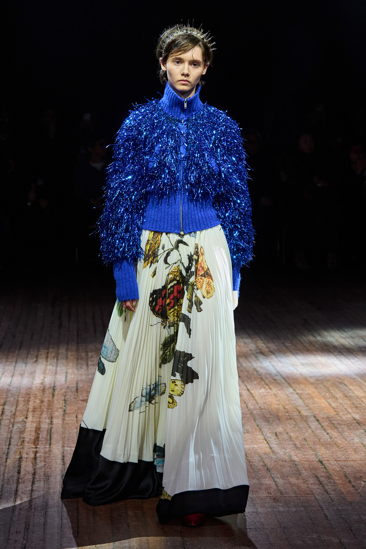 Model on a runway wearing a bright blue, fluffy fringe jacket, a blue turtleneck, and a pleated cream skirt with botanical prints, plus a silver spiked crown.
