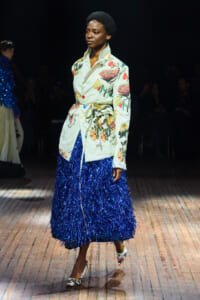 Model walking a runway in a pale floral trench with a bright blue fringe skirt and patterned heels.