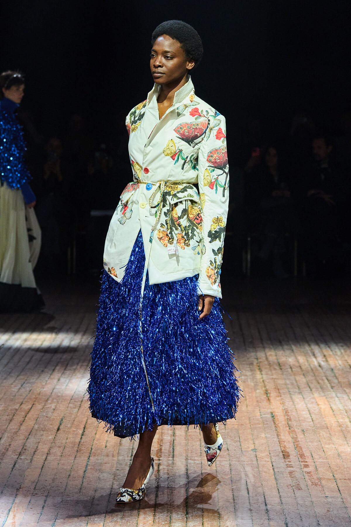 Model walking a runway in a pale floral trench with a bright blue fringe skirt and patterned heels.