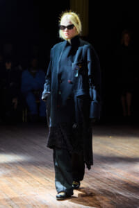 Model on a runway wearing a dark oversized coat with a star badge, spiked halo headpiece, and sunglasses.